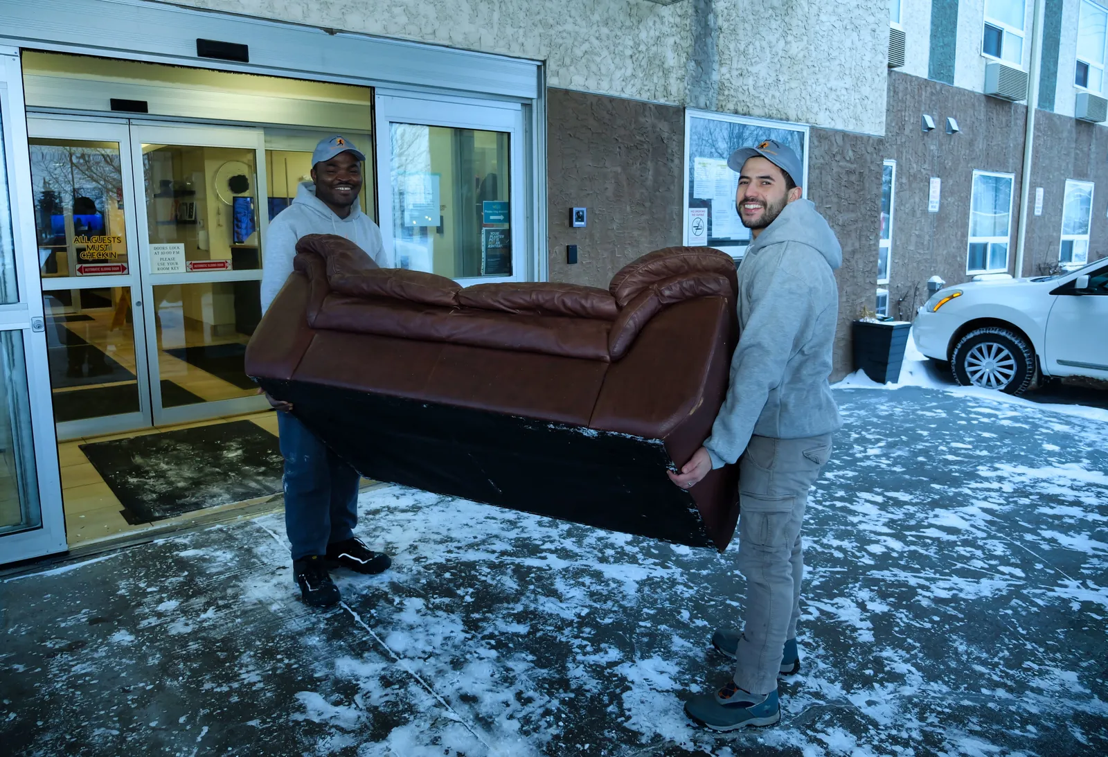 Two Sunshine Junk Removal crew members carrying a brown leather couch out of an apartment entrance in winter